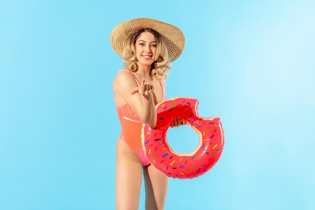 Portrait d'une jolie femme souriante en maillot de bain et chapeau de soleil tenant un anneau en caoutchouc faisant un geste invitant à la station Repos et amusement sur la plage d'été studio tourné isolé sur fond bleu