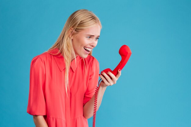 Portrait d'une jolie femme souriante en robe rouge regardant un tube de téléphone rouge isolé sur un mur bleu