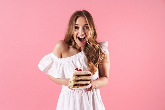 Portrait D'une Jolie Femme Surprise Portant Une Robe Blanche Souriant à La Caméra Et Tenant Un Morceau De Gâteau Avec Une Bougie Isolée Sur Un Mur Rose