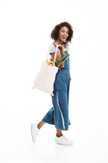 Portrait d'une jolie femme tenant un sac en coton avec des produits alimentaires et marchant isolée sur un mur blanc