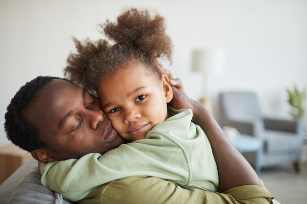 Portrait d'une jolie fille afro-américaine embrassant son père et regardant la caméra dans un intérieur confortable, espace pour copie