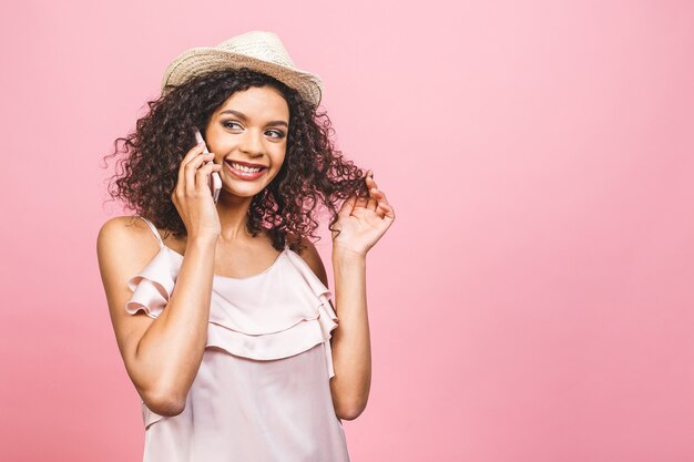 Portrait d'une jolie fille afro-américaine heureuse en robe parlant au téléphone mobile et riant isolé sur fond rose.