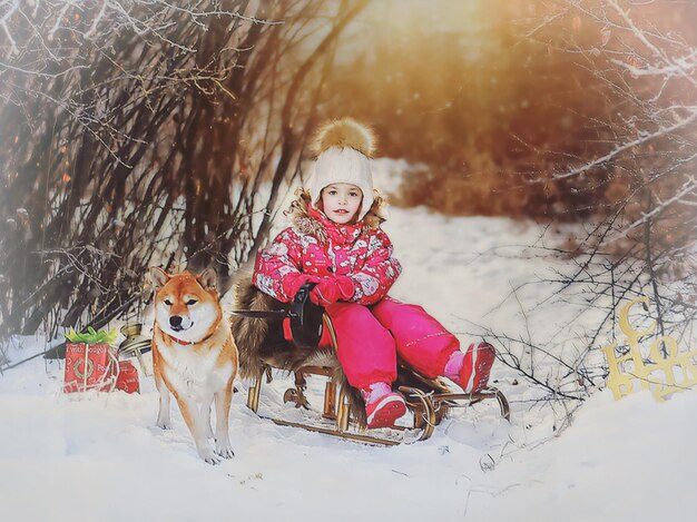 Photo portrait d'une jolie fille assise sur un traîneau par un chien dans une forêt couverte de neige