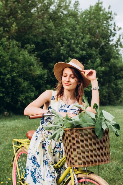 Portrait de jolie fille aux cheveux blonds bouclés en chapeau conduisant un vélo. Des gens actifs. En plein air. Belle fille en vélo dans la campagne, sentant les fleurs, mode de vie d'été. portrait agrandi