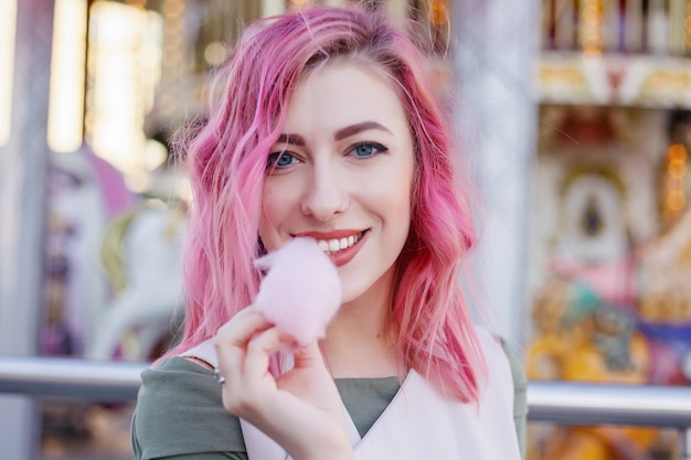 Portrait de jolie fille aux cheveux roses avec une coupe courte posant dans un parc d'attractions sur fond de carrousel. femme aux cheveux ombrés roses avec des paillettes phototeintant des boucles de cheveux à la mode brillante