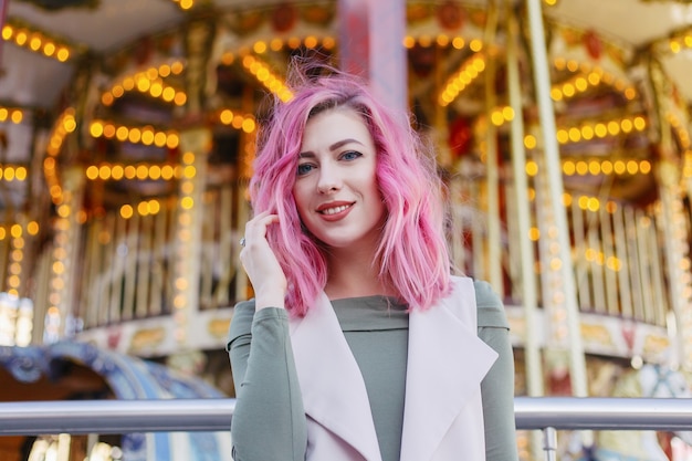 Portrait de jolie fille aux cheveux roses avec une coupe courte posant dans un parc d'attractions sur fond de carrousel. femme aux cheveux ombrés roses avec des paillettes phototeintant des boucles de cheveux à la mode brillante