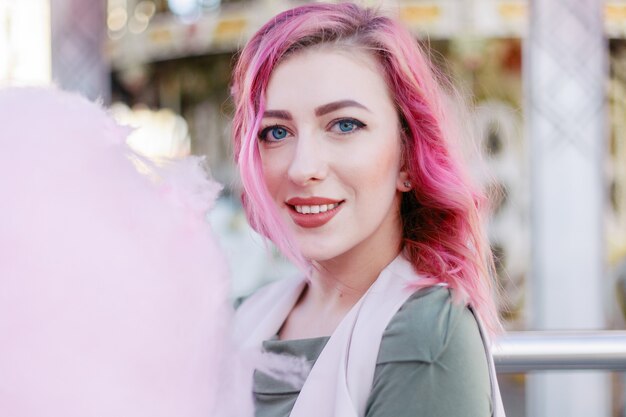 Portrait de jolie fille aux cheveux roses avec une coupe courte posant dans un parc d'attractions sur fond de carrousel. Femme aux cheveux rose avec des paillettes, des boucles de cheveux photodyeing, des paillettes brillantes