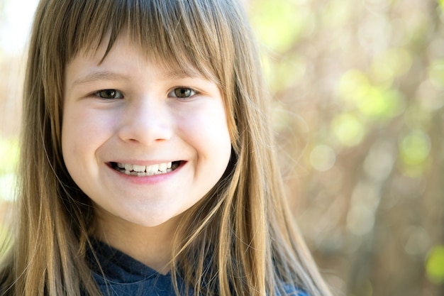 Portrait de jolie fille enfant aux yeux gris et longs cheveux blonds souriant à l'extérieur sur une surface lumineuse floue