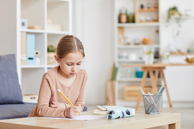 Portrait de jolie fille à faire ses devoirs pour l'école primaire tout en étudiant à la maison dans un intérieur confortable, copiez l'espace
