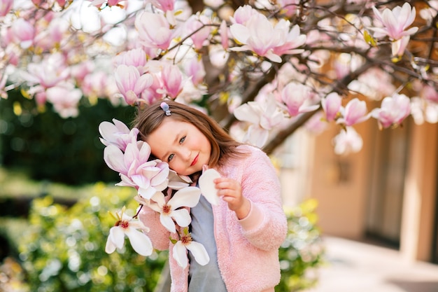 Portrait d'une jolie fille gaie dans une robe grise portant une veste Rosa séjournant au printemps dans un parc sous un magnolia rose en fleurs.