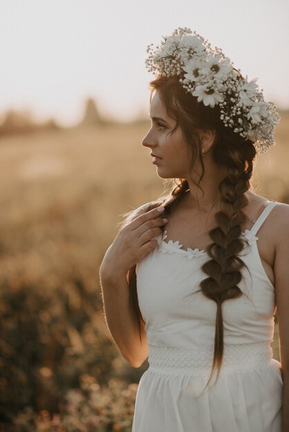 Photo portrait de jolie fille avec guirlande de fleurs et tresses en robe blanche dans le champ de l'été au coucher du soleil