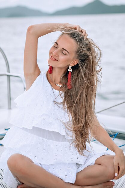 Portrait d'une jolie fille heureuse avec une robe blanche, de longues boucles d'oreilles rouges et de longs cheveux blonds bouclés debout sur un yacht en été. regardant la caméra avec un sourire à pleines dents.