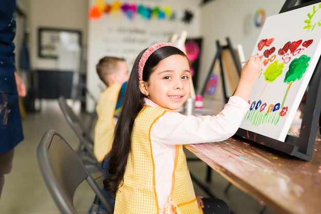 Portrait d'une jolie fille hispanique avec une expression heureuse tout en peignant un paysage sur une toile pour sa classe d'art