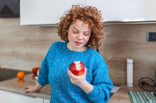 Portrait d'une jolie fille jolie souriante mord une pomme dans sa cuisine. Jeune femme appréciant son fruit de pomme biologique rouge. Apport quotidien de vitamines avec fruits, alimentation et alimentation saine