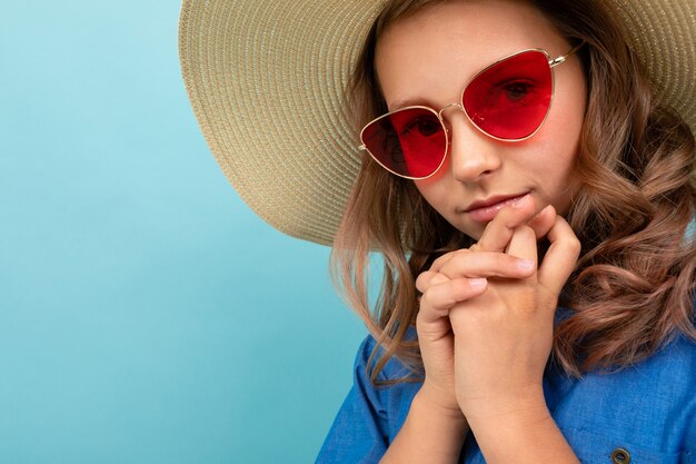 Portrait de jolie fille modèle aux cheveux châtains ondulés en robe, grand chapeau, lunettes de soleil rouges sourit isolé sur fond bleu