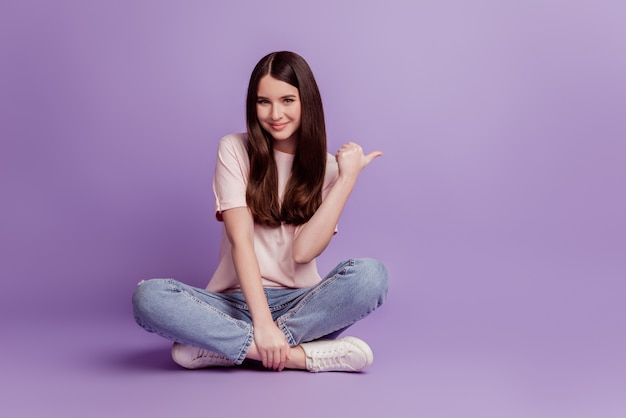 Portrait D'une Jolie Fille Pointant Le Doigt Sur Le Sol Assis Sur Un Mur Violet