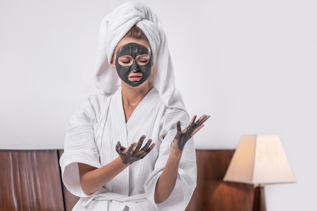 Portrait d'une jolie fille posant dans un masque d'argile avec des mains d'argile dans un peignoir blanc et une serviette sur la tête posant en regardant vers le bas. Concept de détente.