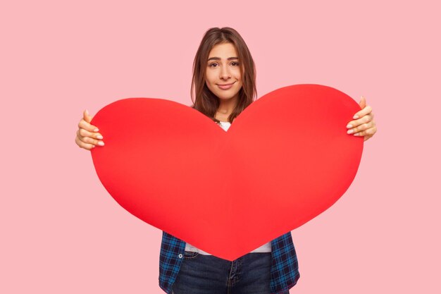 Portrait d'une jolie fille positive tenant un grand coeur en papier rouge et regardant la caméra avec un sourire aimable montrant le symbole de l'amour, des soins et de la charité, prise de vue en studio à l'intérieur isolée sur fond rose