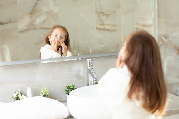 Portrait d'une jolie fille qui se regarde dans le miroir en blouse blanche avec une licorne dans une salle de bain lumineuse avec fenêtre. Hygiène. Routine matinale. Pureté. Comme mère. photo de haute qualité