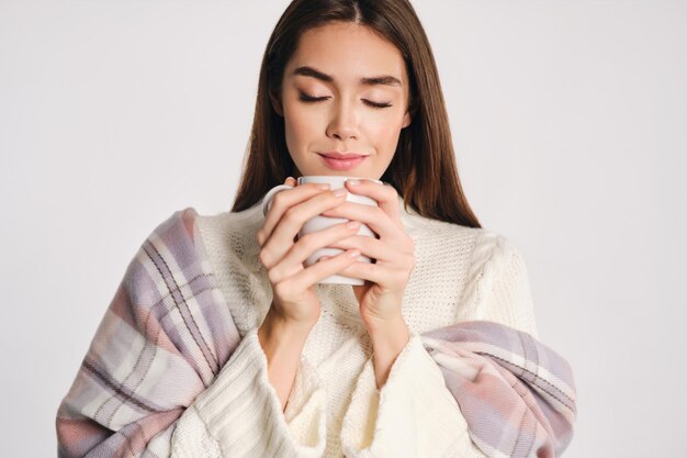 Portrait de jolie fille rêveuse en pull confortable avec plaid sur les épaules tenant une tasse avec une boisson chaude sur fond blanc