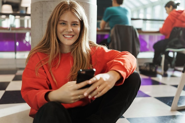 Portrait d'une jolie fille rousse souriante et heureuse assise sur le sol d'un café ou d'un espace de coworking en utilisant des SMS sur un téléphone portable ou en regardant un webinaire en ligne en direct s'auto-éduquer avec une application utile