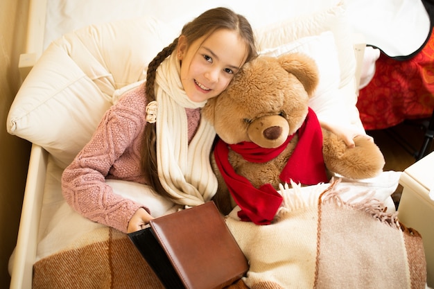 Portrait d'une jolie fille souriante lisant un livre avec un ours en peluche au lit
