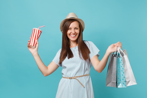 Portrait jolie fille souriante à la mode en robe d'été, chapeau de paille tenant des sacs de paquets avec des achats après le shopping, tasse d'eau gazeuse isolée sur fond bleu pastel. Espace de copie pour la publicité