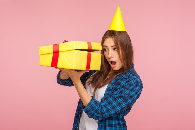 Portrait d'une jolie fille surprise curieuse avec un cône de fête sur la tête regardant à l'intérieur de la boîte cadeau avec une expression étonnée et choquée, étonnée par le cadeau d'anniversaire. studio d'intérieur tourné isolé sur fond rose