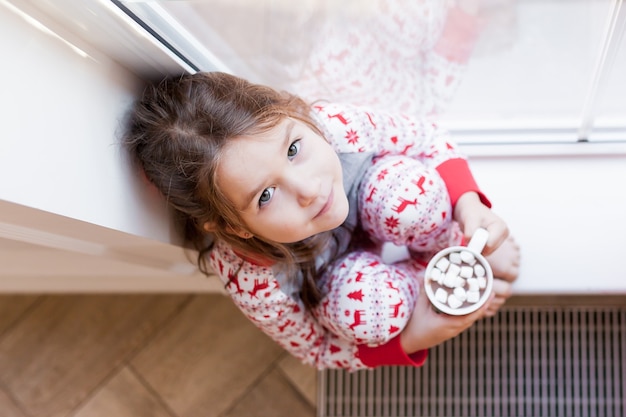 Portrait De Jolie Fille Tenant Du Chocolat Chaud Assis Par Windows. Moments de Noël avec des enfants à la maison