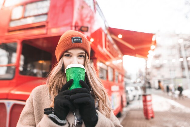 Portrait d'une jolie fille sur des vêtements chauds, debout sur le fond d'un café bus rouge, buvant du café dans une tasse