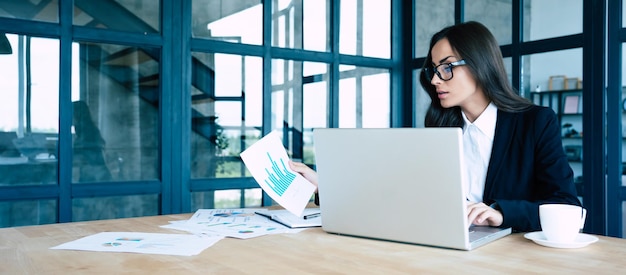 Portrait d'une jolie jeune femme d'affaires souriante dans des verres et un costume complet assis sur le lieu de travail au bureau