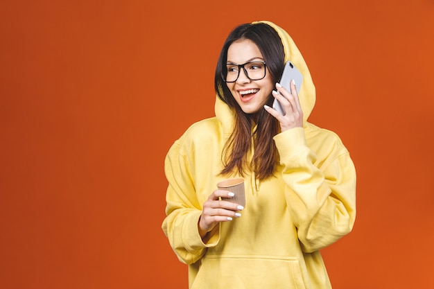 Portrait d'une jolie jeune femme à l'aide de téléphone portable tout en tenant une tasse de café à emporter isolé sur fond orange.