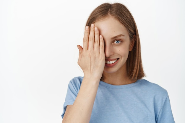 Portrait d'une jolie jeune femme aux cheveux blonds courts, couvrant la moitié du visage, regardant d'un œil et souriant à la caméra, soins de la peau avant après concept, fond blanc