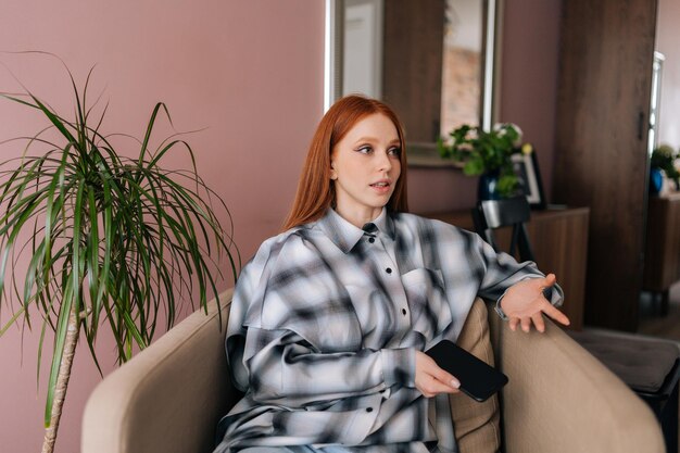 Portrait d'une jolie jeune femme aux cheveux roux à l'aide d'un smartphone assis sur un fauteuil moelleux à la maison en détournant les yeux