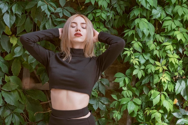 Portrait de jolie jeune femme aux yeux fermés cheveux blonds en haut court noir sur fond de plantes vertes