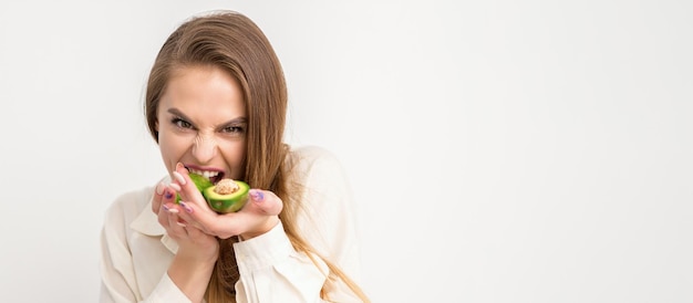 Portrait d'une jolie jeune femme caucasienne brune souriante portant la chemise blanche aux cheveux longs tenant et montrant l'avocat debout isolé sur fond blanc