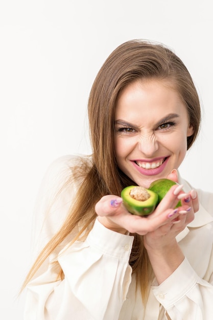 Portrait d'une jolie jeune femme caucasienne brune souriante portant la chemise blanche aux cheveux longs tenant et montrant l'avocat debout isolé sur fond blanc