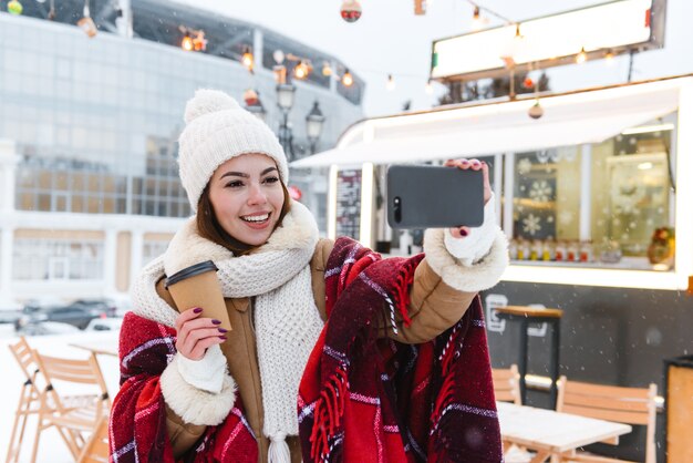 Portrait d'une jolie jeune femme en chapeau et écharpe marchant à l'extérieur en hiver neige boire du café prendre un selfie par téléphone mobile.