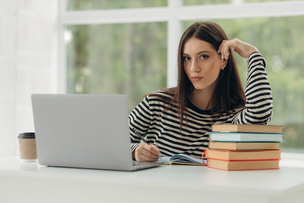 Portrait d'une jolie jeune femme étudiant assis à table avec un ordinateur portable et des livres à la maison Concept d'apprentissage