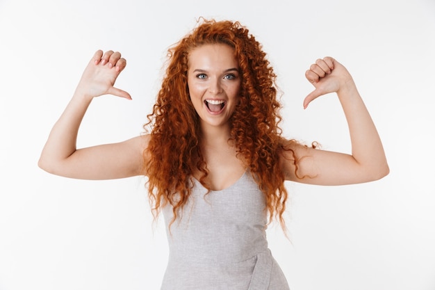 Portrait d'une jolie jeune femme gaie avec de longs cheveux roux bouclés isolés, pointant sur elle-même