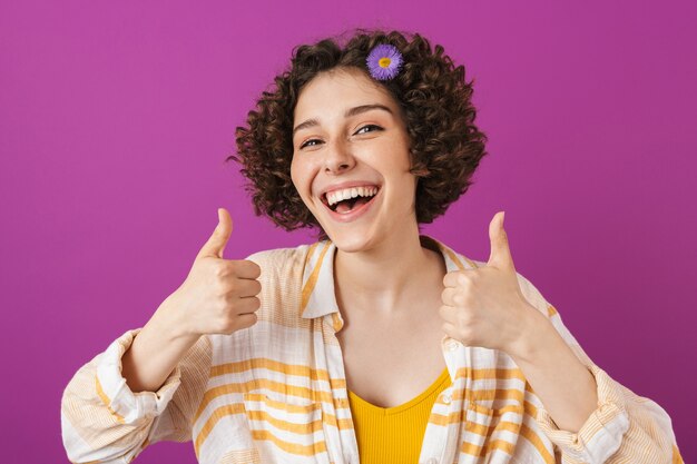 Portrait d'une jolie jeune femme heureuse avec des cheveux bruns bouclés debout isolé sur mur violet, pouces vers le haut