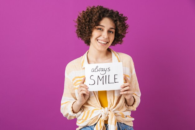 Portrait d'une jolie jeune femme heureuse avec des cheveux bruns bouclés isolés sur un mur violet, tenant une planche avec des lettres de motivation