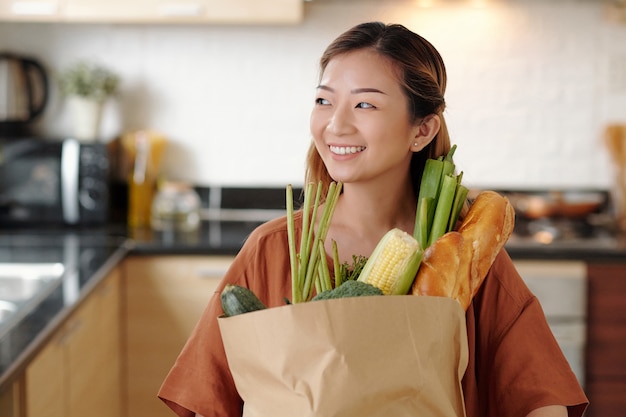 Portrait d'une jolie jeune femme heureuse debout dans la cuisine avec un sac en papier d'épicerie fraîche comme du pain et des légumes