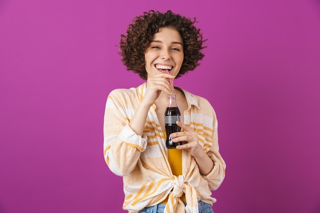 Portrait d'une jolie jeune femme joyeuse aux cheveux bouclés brune debout isolée sur un mur violet, tenant une bouteille en verre avec une boisson gazeuse