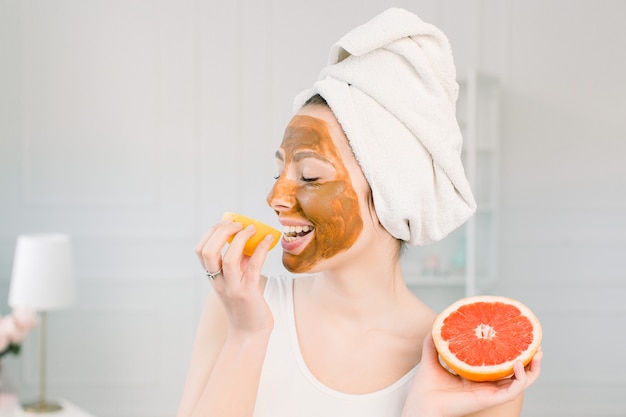 Portrait de jolie jeune femme avec un masque à l'argile sur son visage tenant des tranches de citron et de pamplemousse, soins du visage