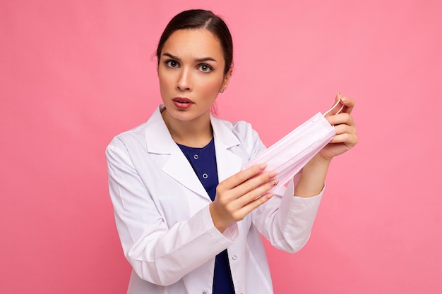 Portrait d'une jolie jeune femme médecin en colère en blouse blanche tenant un masque médical