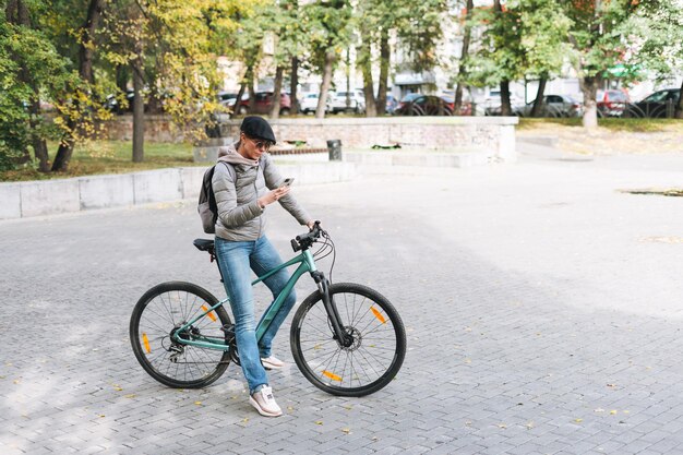 Portrait d'une jolie jeune femme à la mode en casquette et lunettes de soleil à l'aide de mobile à vélo le jour d'automne ensoleillé dans le parc de la ville