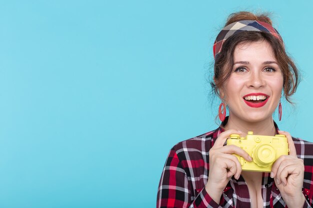 Portrait d'une jolie jeune femme positive dans une chemise à carreaux tenant un appareil photo vintage de film jaune posant sur un mur bleu. Amateurs de concept de photographie et de prise de vue. espace promo