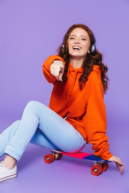 Portrait d'une jolie jeune femme rousse assise sur une planche à roulettes sur violet, écoutant de la musique avec des écouteurs