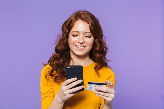Portrait d'une jolie jeune femme rousse joyeuse debout sur violet, à l'aide de téléphone mobile, montrant la carte de crédit